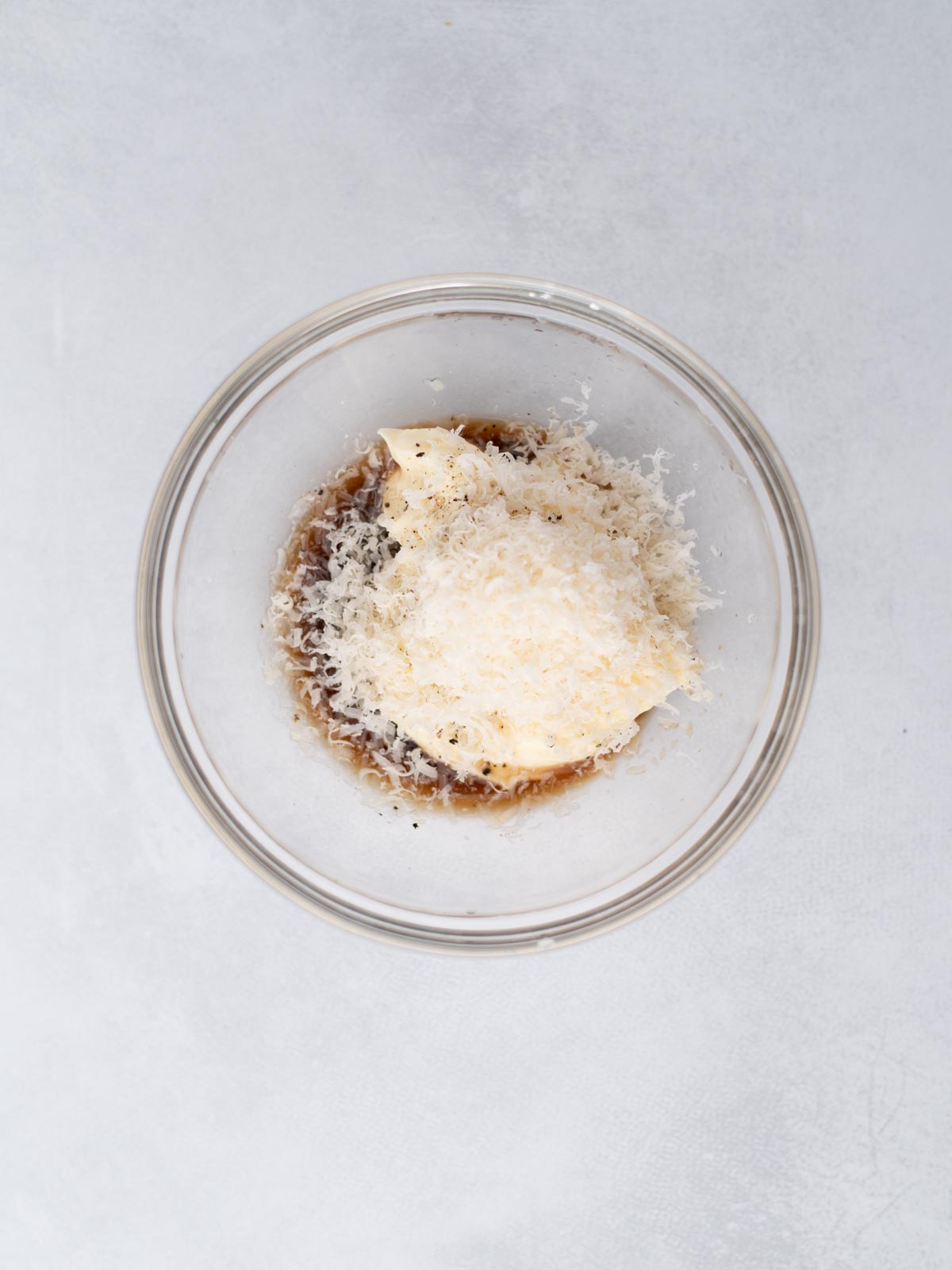 A glass bowl containing grated cheese, mayonnaise, Worcestershire sauce, and black pepper, set on a light gray surface.