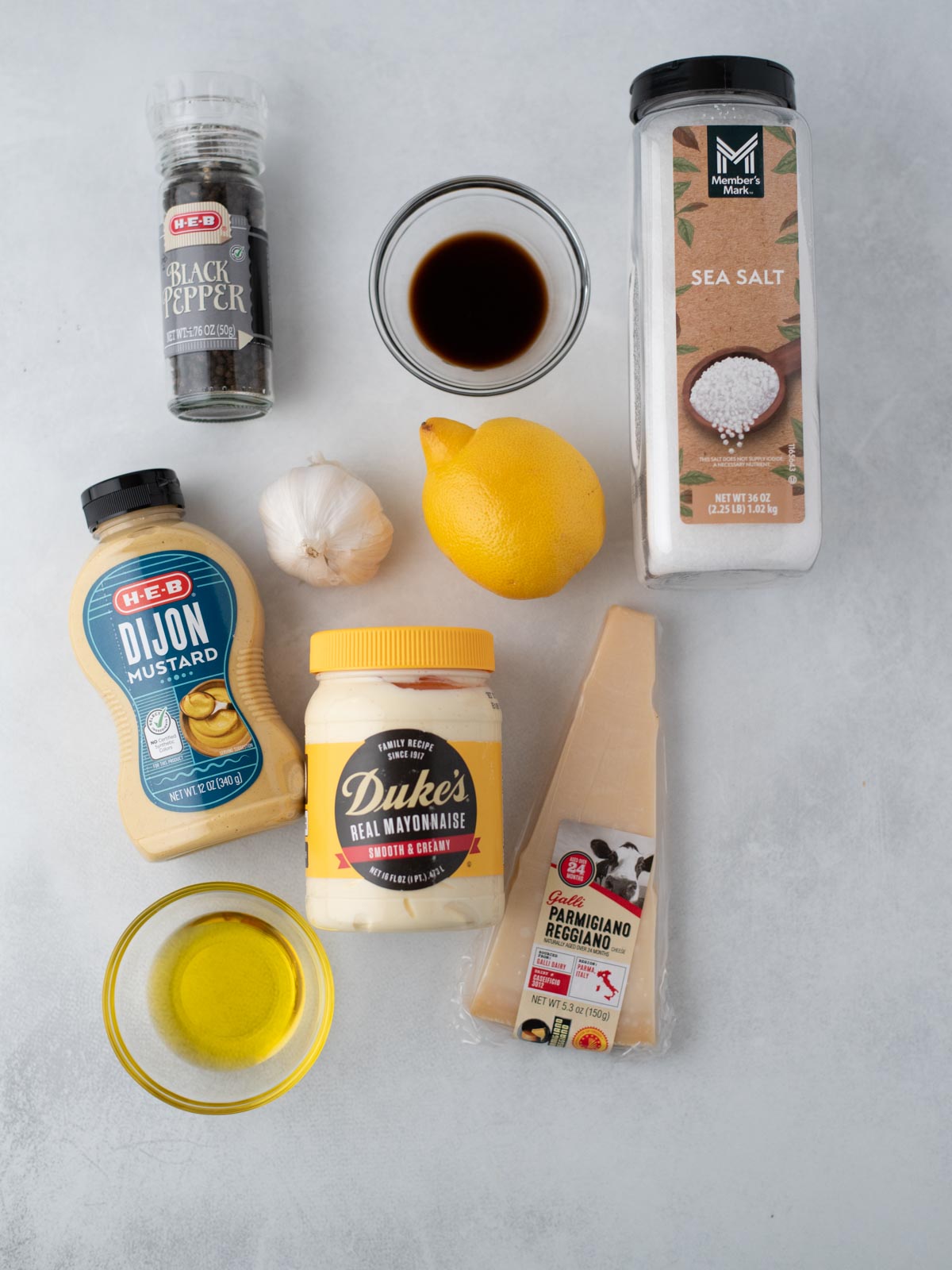 Ingredients for Caesar dressing displayed on a light countertop, including black pepper, Worcestershire sauce, sea salt, Dijon mustard, mayonnaise, a lemon, garlic bulbs, olive oil, and a wedge of Parmesan cheese.