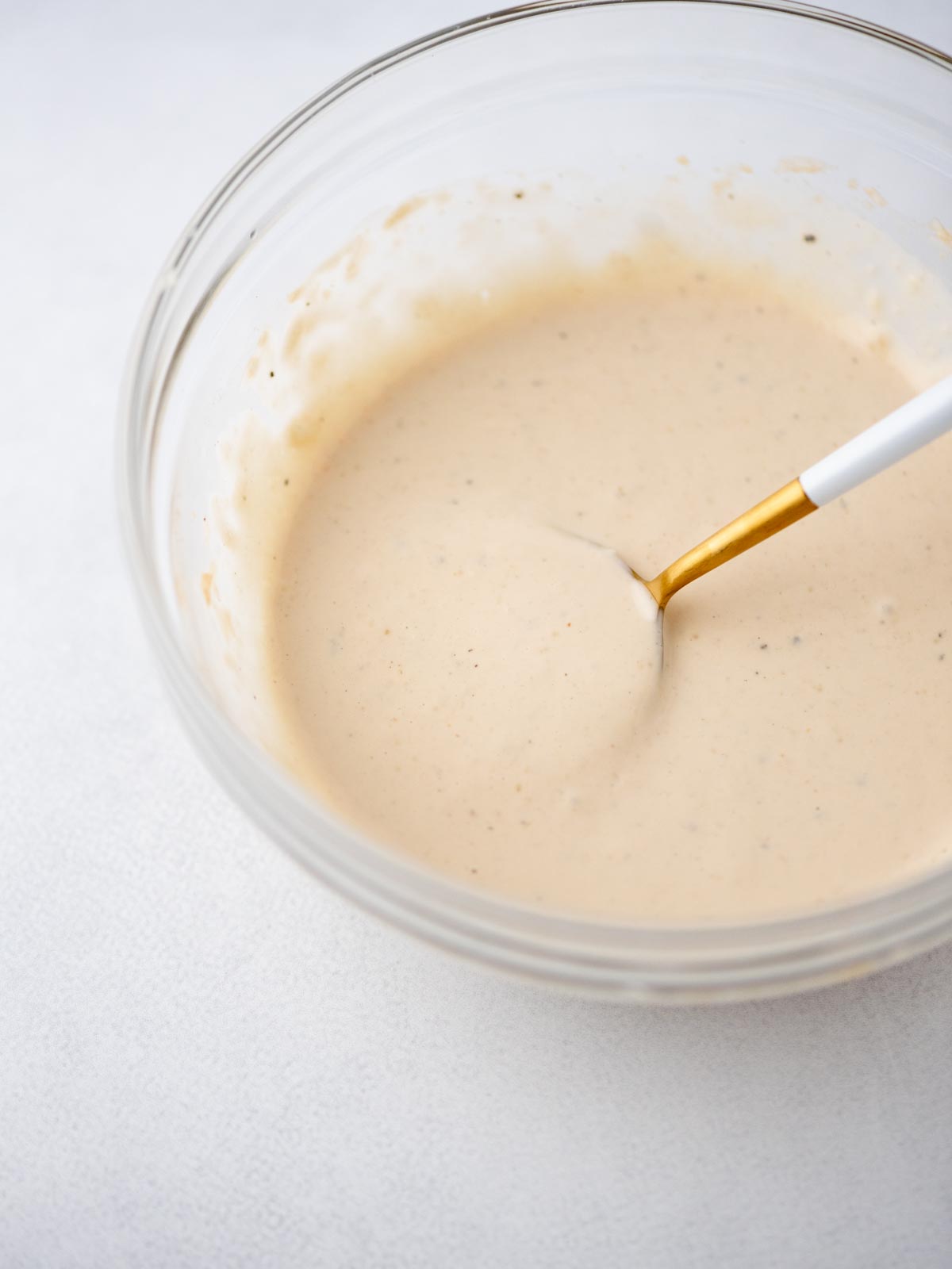A glass bowl filled with creamy, light beige sauce or dressing, being mixed with a white and gold spoon on a light surface.