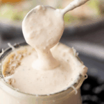 A spoon drips creamy homemade Caesar dressing into a glass jar, with a blurred salad in the background. Text above reads, homemade caesar dressing and sweetlysplendid.com.