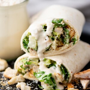 A close-up of a chicken Caesar salad wrap cut in half, filled with lettuce, chicken, croutons, and creamy dressing, with sauce drizzling out and crouton pieces scattered on the plate.