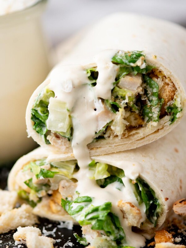 A close-up of a chicken Caesar salad wrap cut in half, filled with lettuce, chicken, croutons, and creamy dressing, with sauce drizzling out and crouton pieces scattered on the plate.