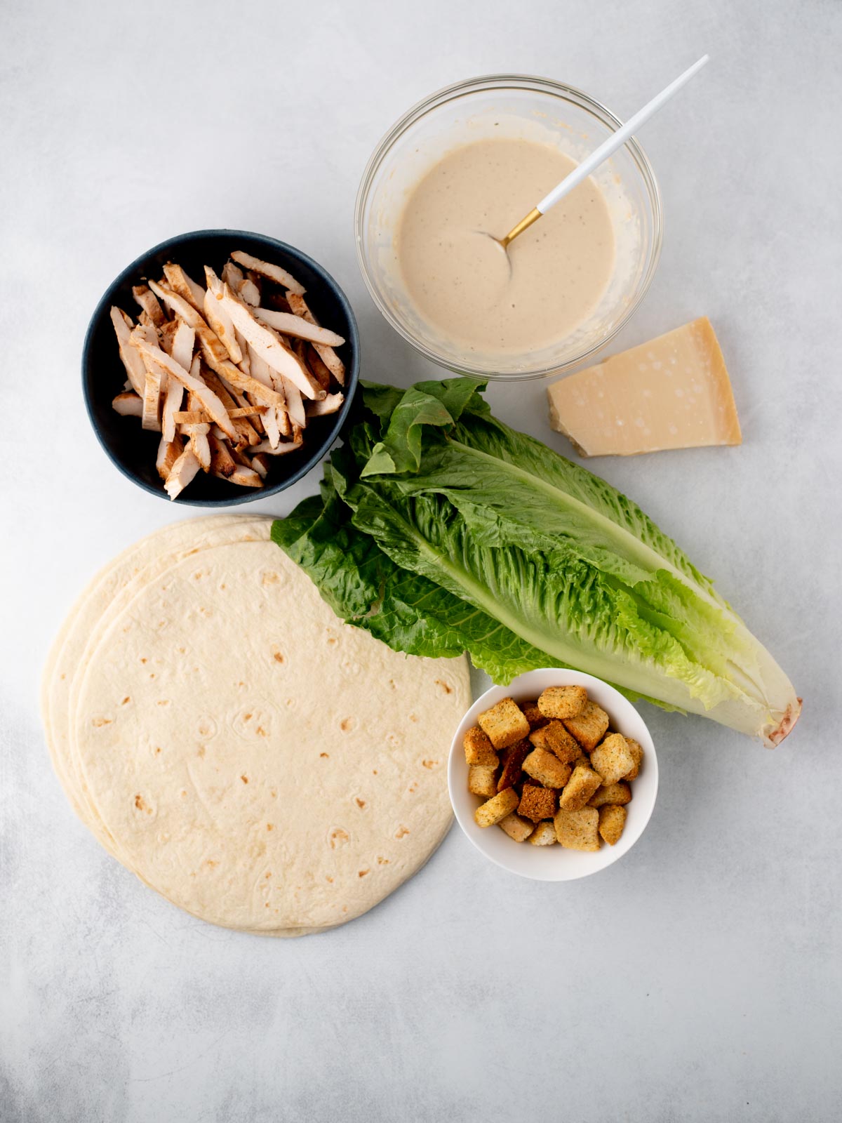 Ingredients for a chicken Caesar wrap: flour tortillas, sliced grilled chicken, romaine lettuce, Caesar dressing in a bowl, Parmesan cheese wedge, and a small bowl of croutons on a white surface.