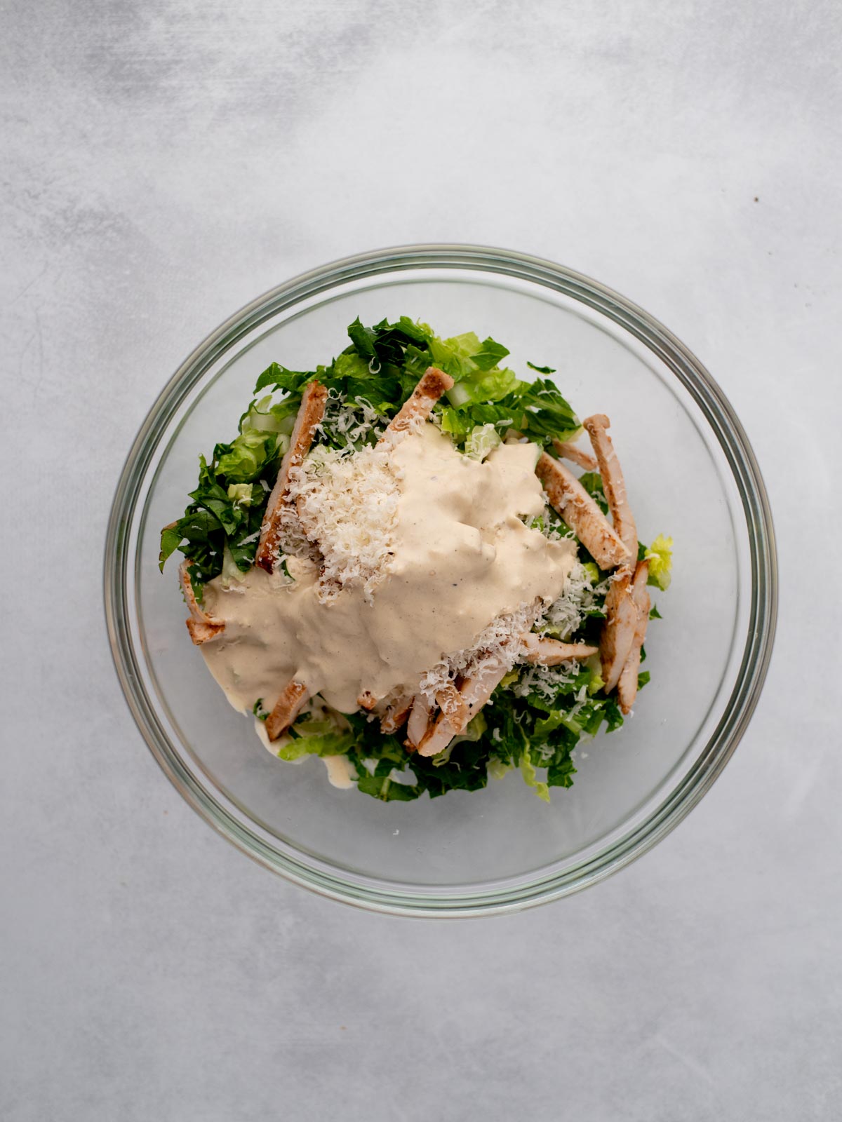 A glass bowl filled with chopped romaine lettuce, grilled chicken strips, grated cheese, and a generous amount of creamy dressing on top, set against a light gray background.