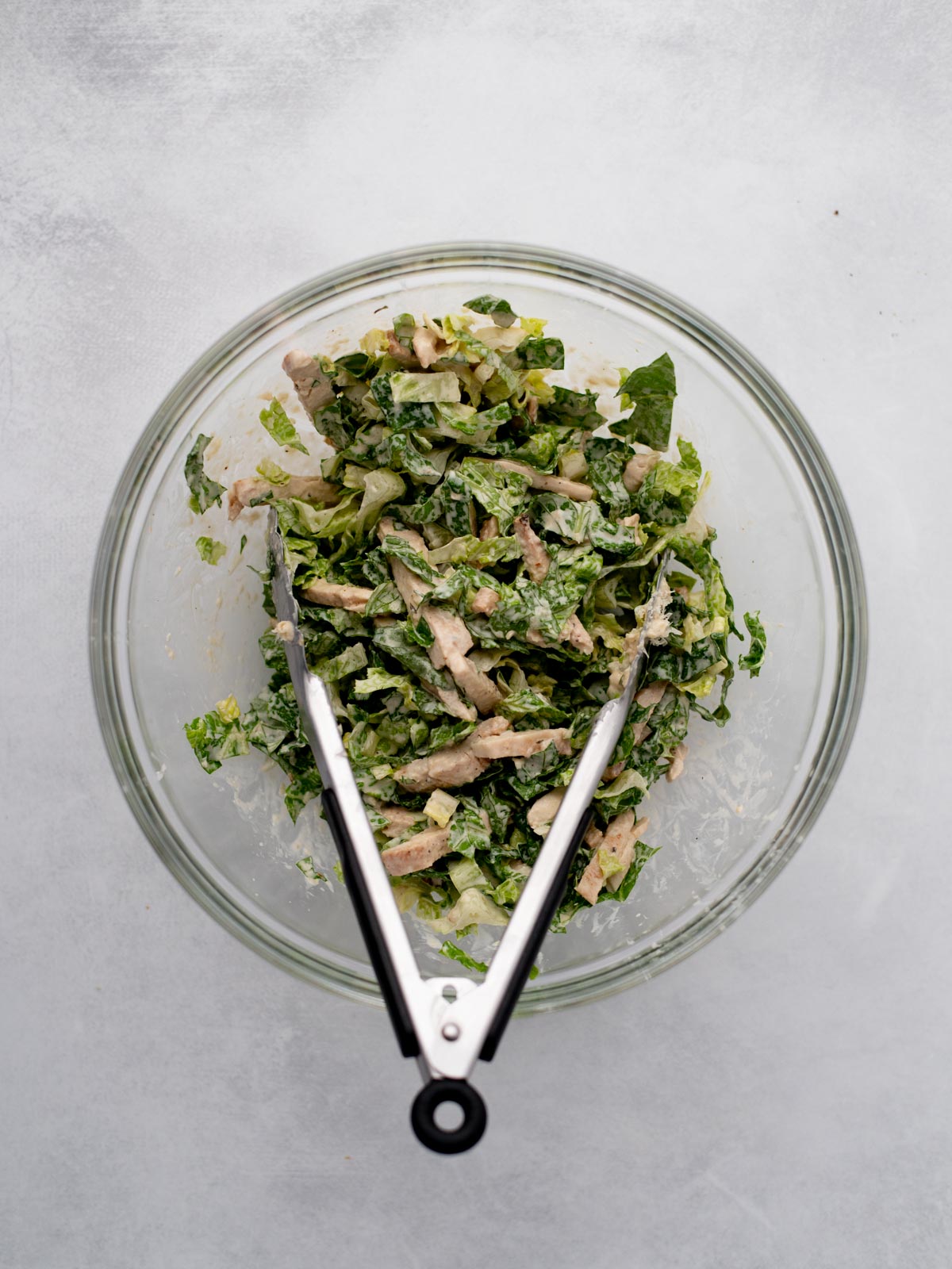 A glass bowl filled with chopped salad greens and pieces of cooked chicken, with metal tongs resting inside the bowl, on a light gray surface.