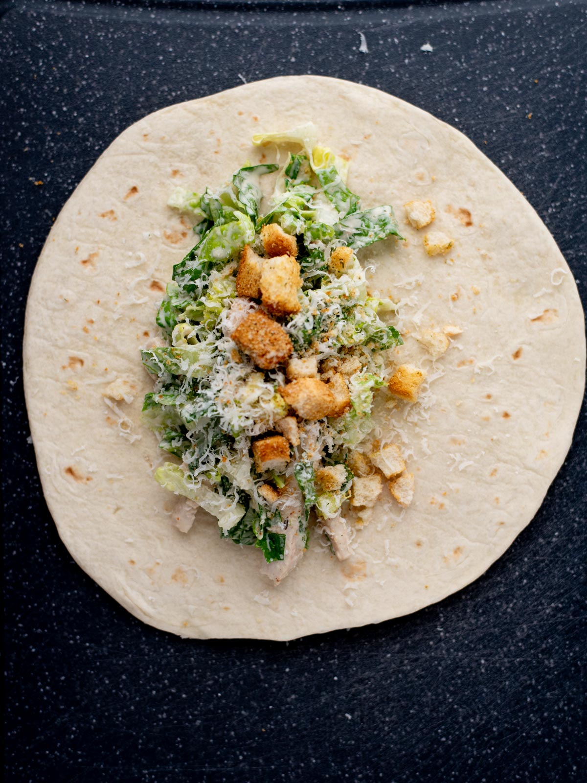 A flour tortilla on a dark surface topped with chopped romaine lettuce, shredded cheese, croutons, and pieces of chicken, ready to be wrapped as a Caesar salad wrap.