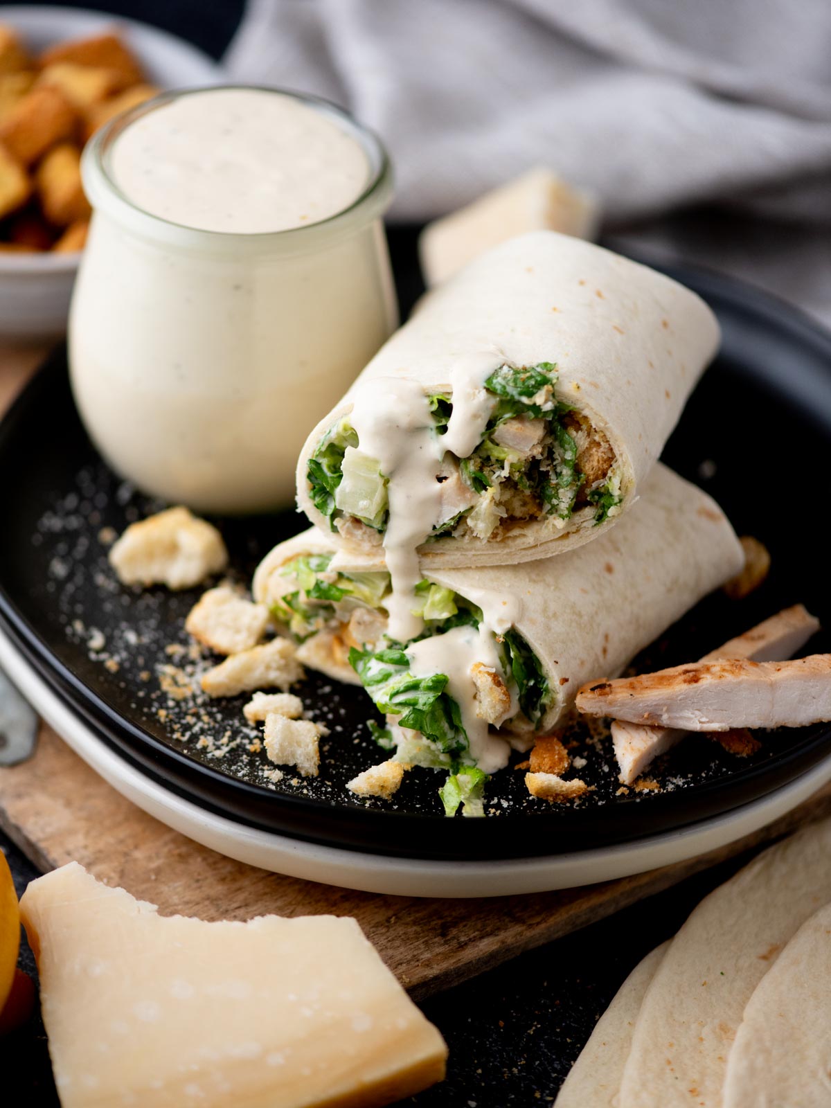 A Caesar salad wrap cut in half sits on a black plate, filled with lettuce, chicken, croutons, and creamy Caesar dressing, with extra dressing in a jar nearby and sprinkled cheese on the plate.