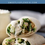 Two chicken Caesar wraps are stacked on a black plate, filled with lettuce, chicken, croutons, and Caesar dressing. A jar of dressing and a piece of grilled chicken are visible in the background.