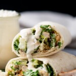 Two chicken Caesar salad wraps are stacked on a plate, filled with lettuce, grilled chicken, croutons, and Caesar dressing, with extra dressing drizzled on top. A jar of dressing and a wedge of cheese are in the background.