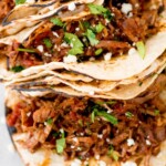 Overhead view of several tacos filled with shredded beef barbacoa, topped with chopped cilantro and crumbled cheese, on toasted tortillas. A label at the top reads slow cooker beef barbacoa - sweetlysplendid.com.