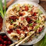 A glass bowl filled with pasta salad featuring bowtie pasta, chopped lettuce, cherry tomatoes, bacon bits, cheese cubes, and green onions, with two serving utensils inside.
