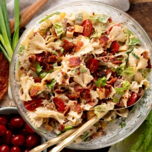 A glass bowl filled with pasta salad featuring bowtie pasta, chopped lettuce, cherry tomatoes, bacon bits, cheese cubes, and green onions, with two serving utensils inside.