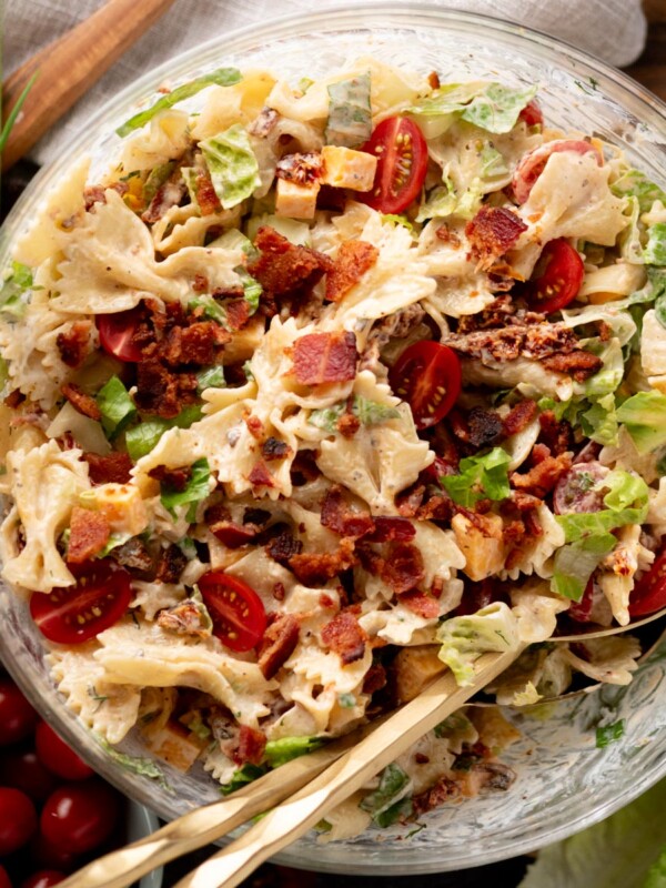 A glass bowl filled with pasta salad featuring bowtie pasta, chopped lettuce, cherry tomatoes, bacon bits, cheese cubes, and green onions, with two serving utensils inside.