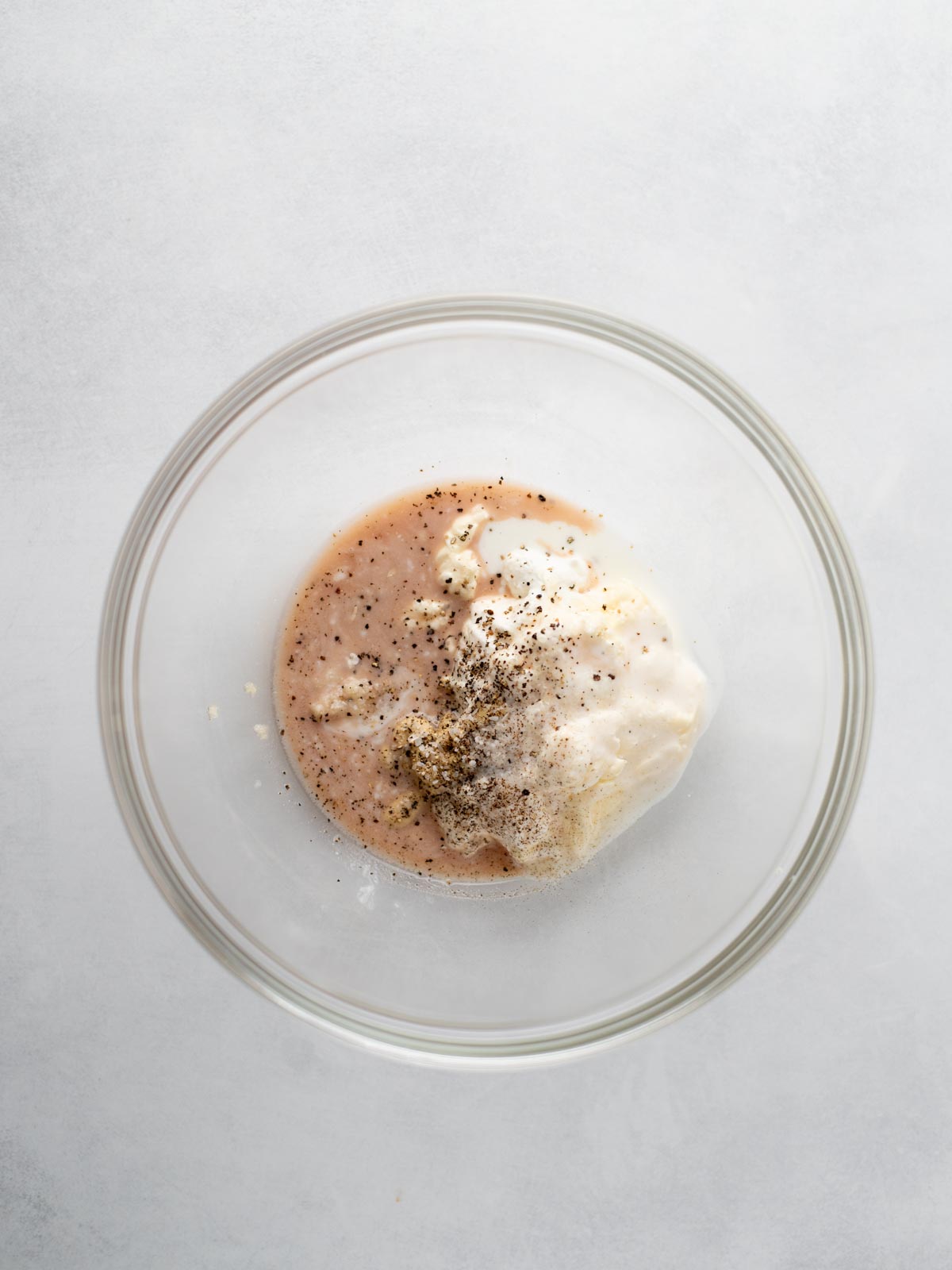 A glass mixing bowl containing mayonnaise, red liquid, and cracked black pepper on a light grey surface, viewed from above.