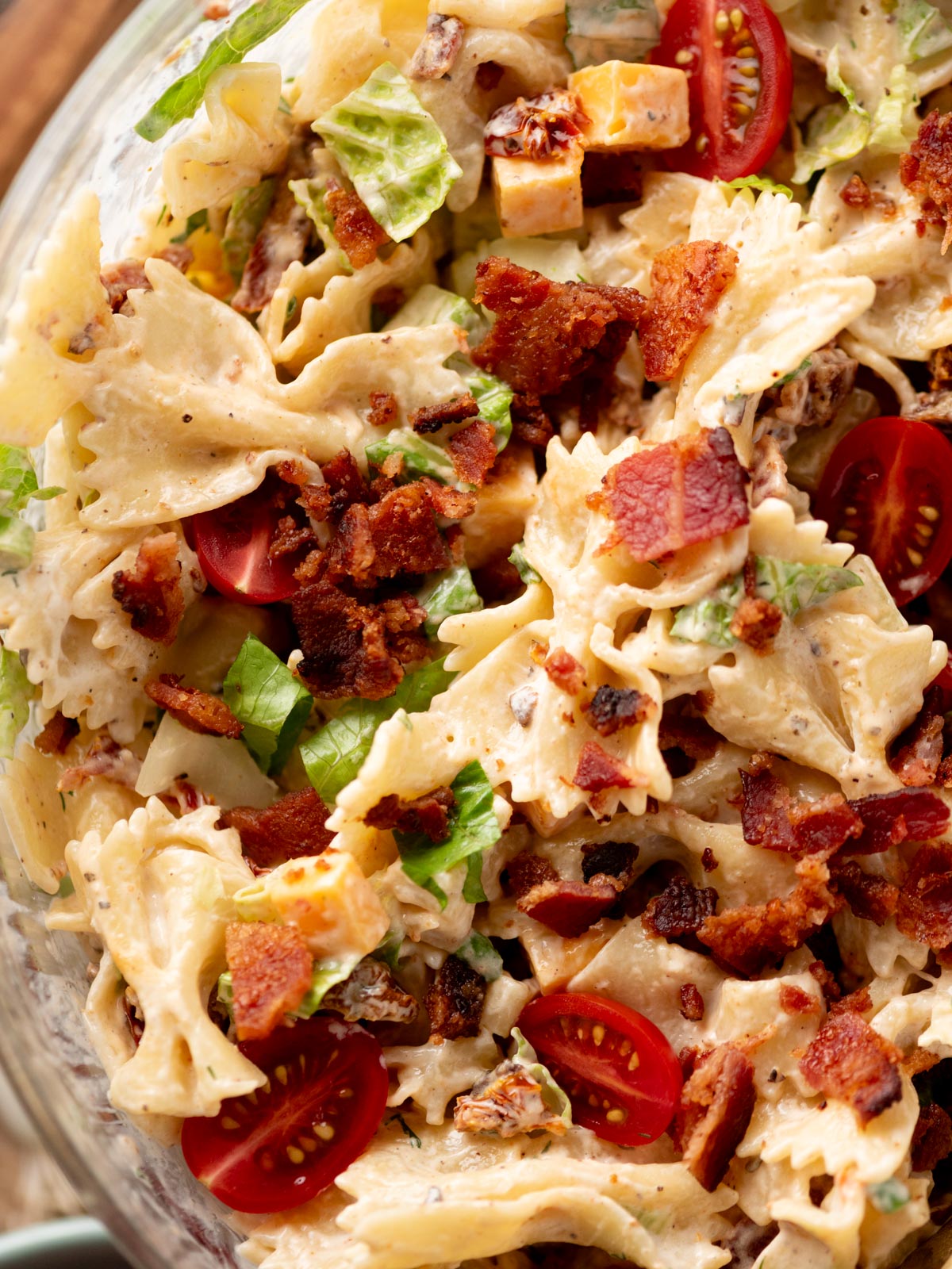 A close-up of a pasta salad with bowtie pasta, cherry tomatoes, lettuce, cubed cheese, bacon pieces, and a creamy dressing.