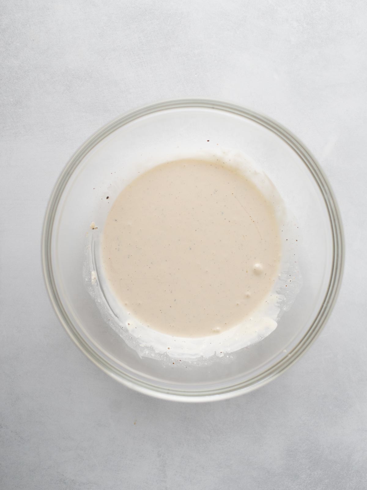 A clear glass bowl containing a small amount of light beige batter or sauce sits on a light gray surface.