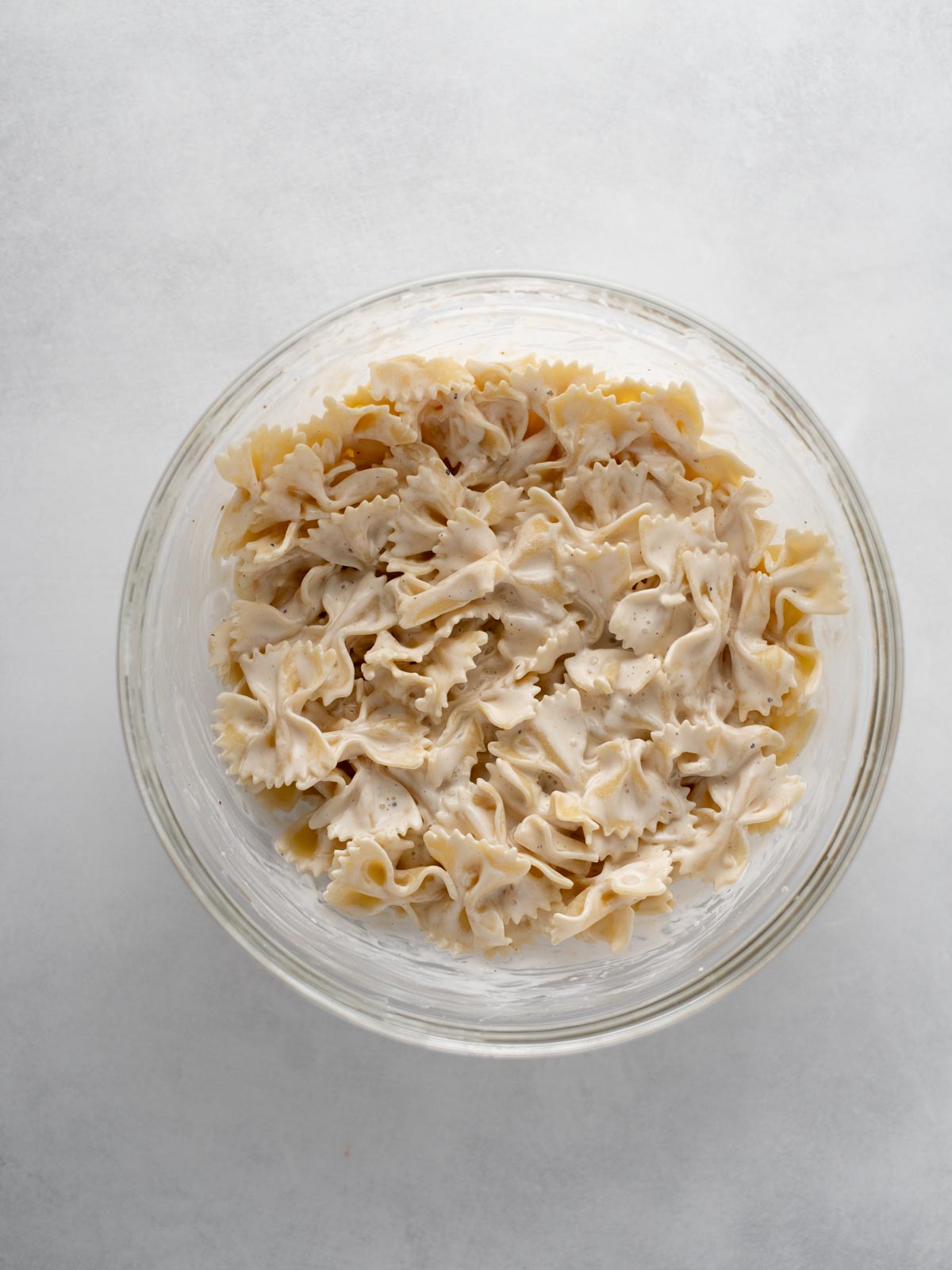 A clear glass bowl filled with farfalle pasta coated in a creamy white sauce, viewed from above on a light gray surface.