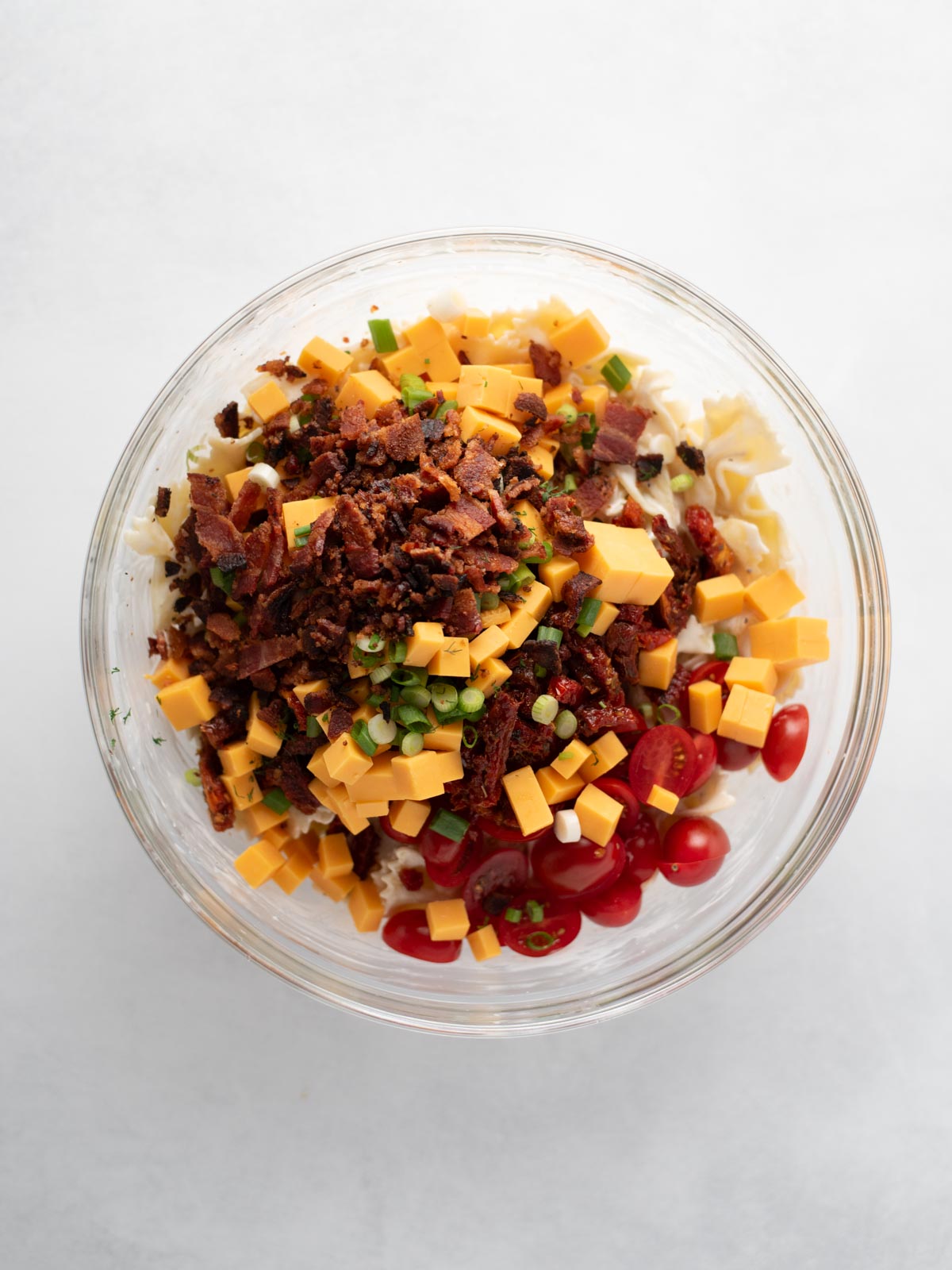 A glass bowl filled with uncooked pasta, diced cheddar cheese, bacon bits, cherry tomatoes, green onions, and herbs, photographed from above on a white background.