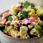 A bowl filled with broccoli salad, featuring broccoli florets, crispy bacon pieces, shredded cheese, chopped red onion, and a creamy dressing. The text above reads creamy bacon broccoli salad.