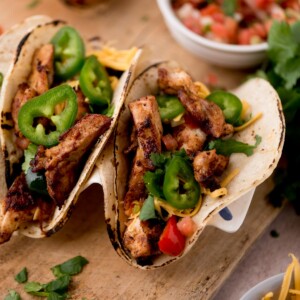 Two grilled chicken tacos on corn tortillas, inspired by easy chicken fajitas, topped with sliced jalapeños, shredded cheese, chopped tomatoes, and cilantro, served on a wooden board with a bowl of salsa in the background.