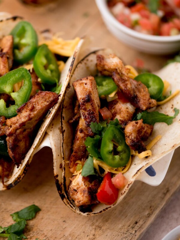 Two grilled chicken tacos on corn tortillas, inspired by easy chicken fajitas, topped with sliced jalapeños, shredded cheese, chopped tomatoes, and cilantro, served on a wooden board with a bowl of salsa in the background.