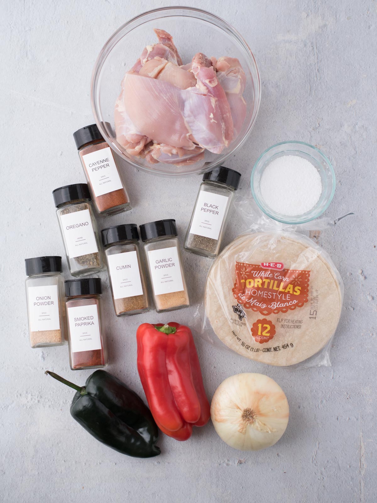 Overhead view of raw chicken in a bowl, a stack of tortillas, salt, red and poblano peppers, onion, and spice jars—perfect ingredients for easy chicken fajitas or classic Texas chicken fajitas.