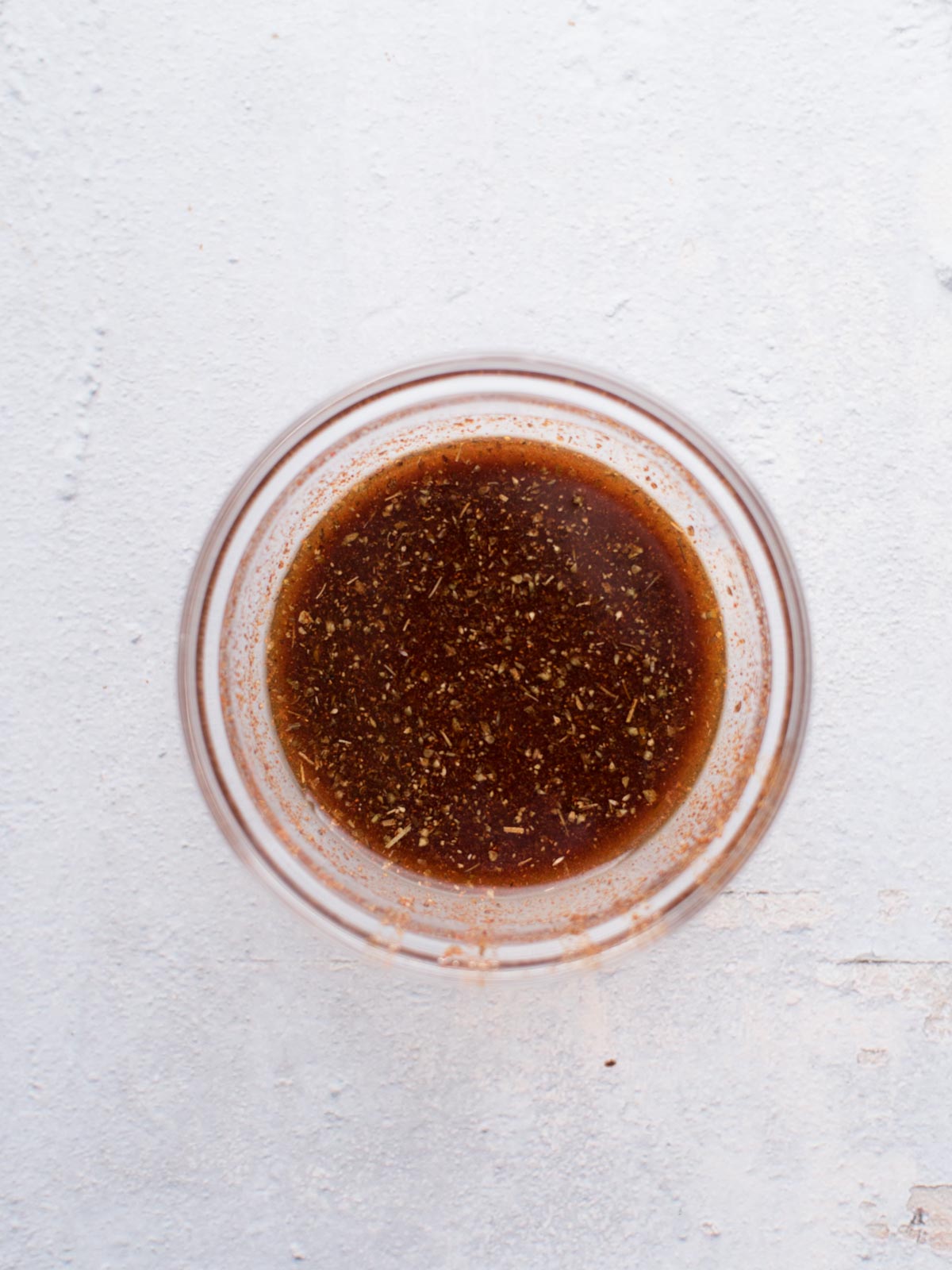 A glass bowl containing a brown, herb-speckled liquid sits on a light, textured surface. This flavorful mixture appears to be a marinade or dressing, perfect for making easy chicken fajitas.