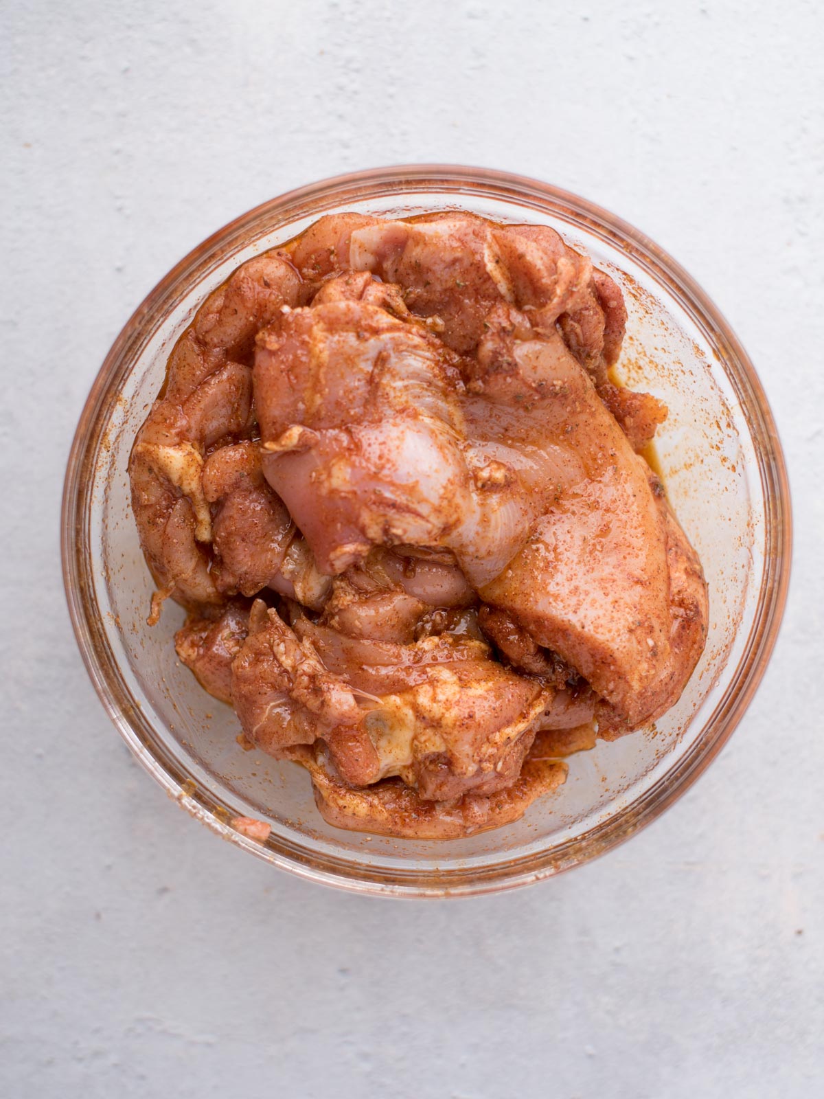A glass bowl filled with raw chicken pieces coated in a reddish-brown marinade, perfect for making easy chicken fajitas, sits on a light-colored surface.