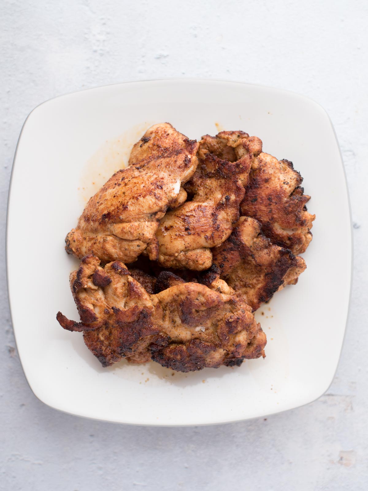 A white plate with several pieces of cooked, seasoned chicken thighs stacked on it, perfect for making easy chicken fajitas, is placed on a light-colored surface.
