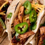 Three Tex-Mex chicken fajitas filled with grilled chicken, shredded cheese, sliced jalapeños, and fresh cilantro on toasted tortillas, displayed on a wooden surface.