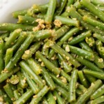 A white bowl filled with garlic green beans coated in garlic butter, topped with minced garlic and seasonings. A label above the bowl reads “garlic butter green beans” with the website sweetlysplendid.com.