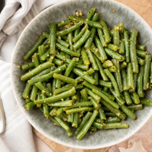 A bowl of cooked green beans cut into pieces, seasoned with herbs and garlic, sits on a wooden surface next to a striped cloth.