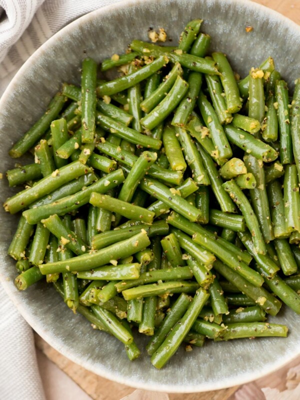 A bowl of cooked green beans cut into pieces, seasoned with herbs and garlic, sits on a wooden surface next to a striped cloth.