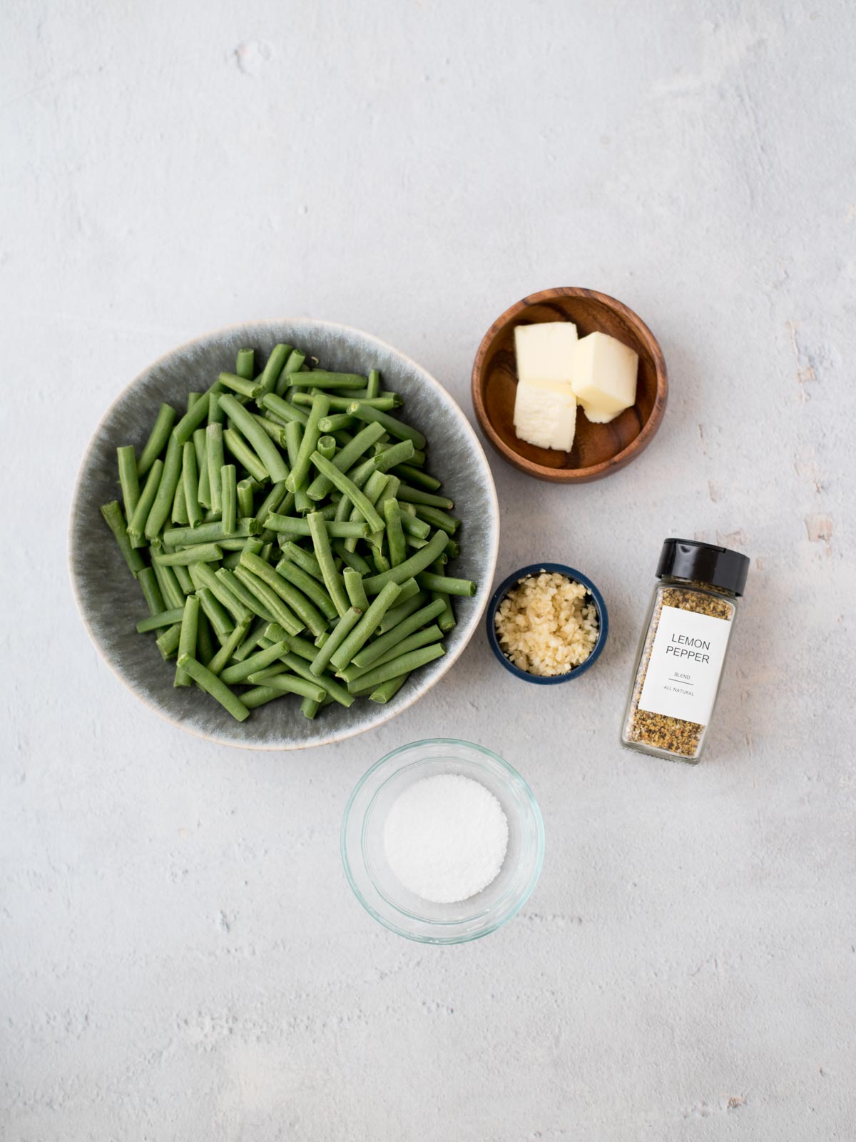 A bowl of cut green beans, a small bowl of butter cubes, a small dish of minced garlic, a jar of lemon pepper seasoning, and a small bowl of salt on a light surface.