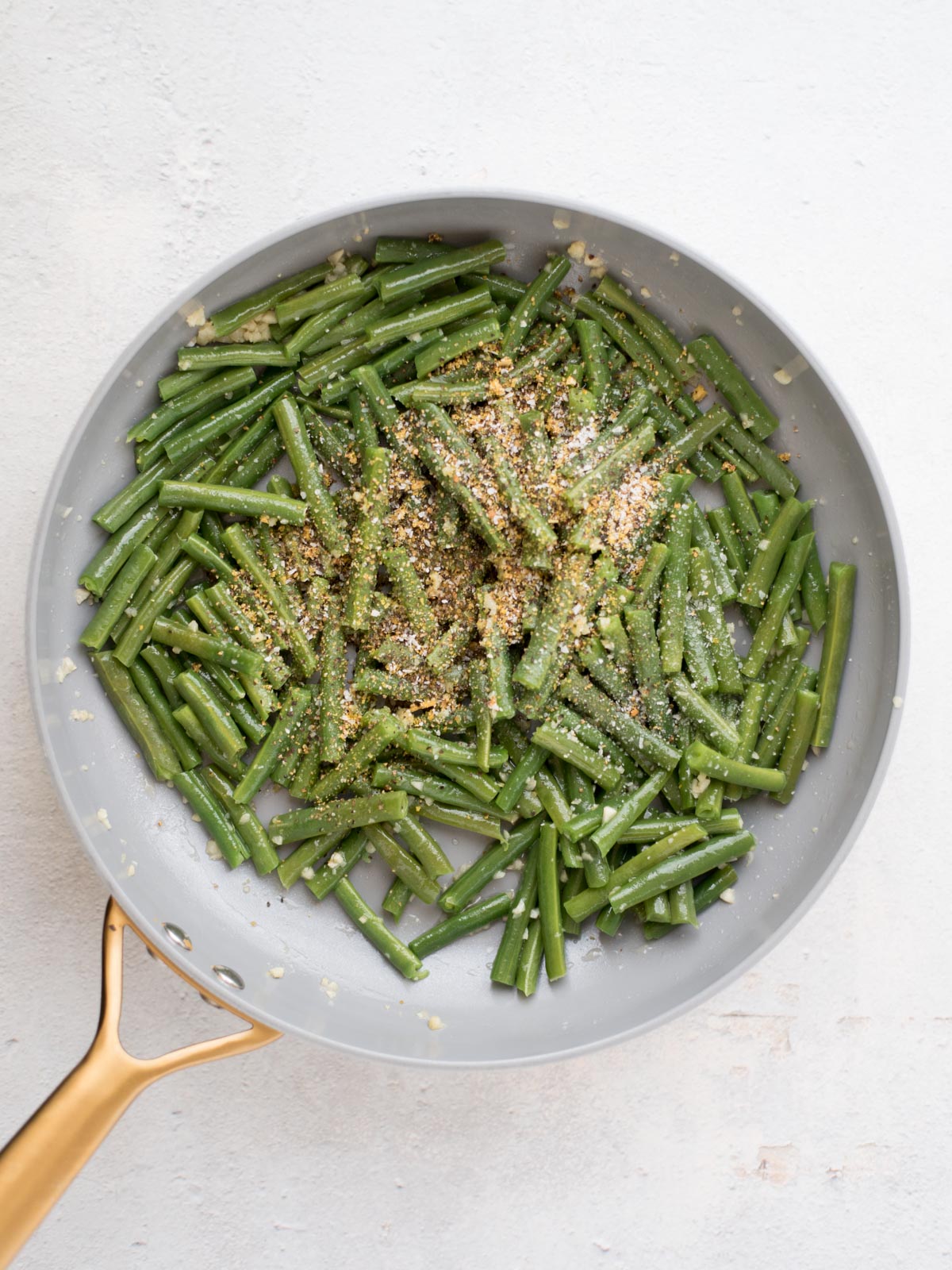 A frying pan with cooked green beans cut into pieces, topped with seasoning and breadcrumbs, on a light-colored surface.