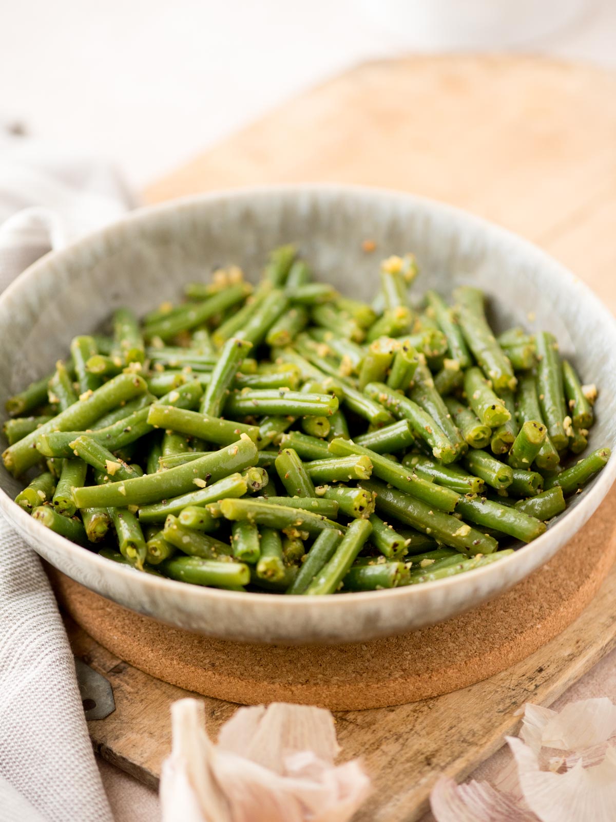 A ceramic bowl filled with cooked green beans tossed with garlic and spices, placed on a wooden surface with garlic cloves nearby.
