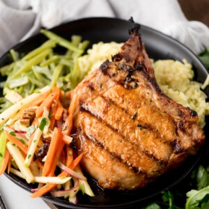 A grilled pork chop takes center stage on a black plate, paired with yellow rice, sautéed green beans, and a fresh apple-carrot slaw tossed with nuts and parsley. Grilled pork chops like this shine beside a light cloth napkin in the background.