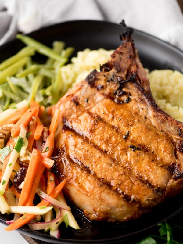 A grilled pork chop takes center stage on a black plate, paired with yellow rice, sautéed green beans, and a fresh apple-carrot slaw tossed with nuts and parsley. Grilled pork chops like this shine beside a light cloth napkin in the background.