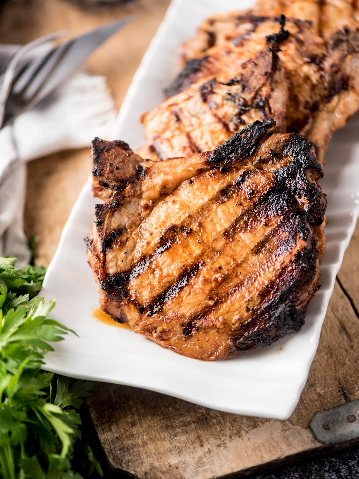 A close-up of juicy grilled pork chops with dark grill marks on a white rectangular plate, garnished with fresh parsley, set on a rustic wooden surface.