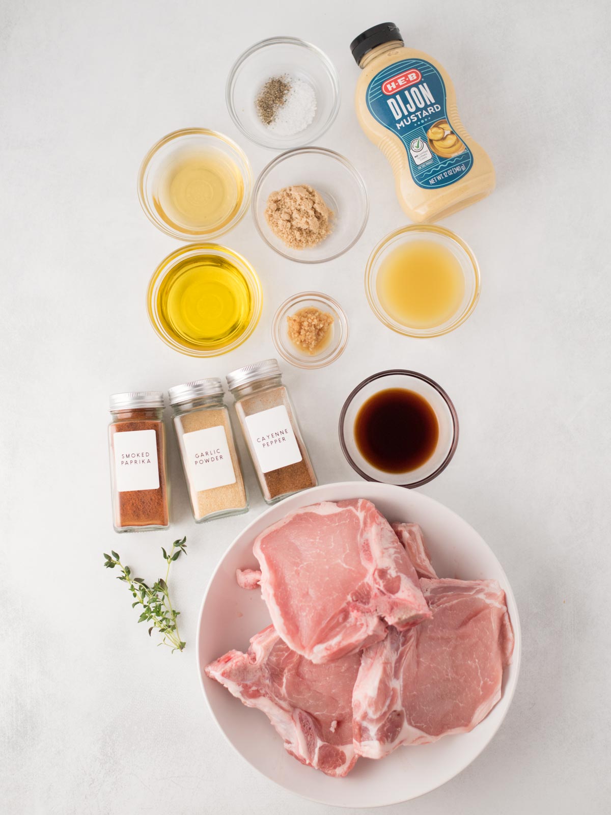 A bowl of raw pork chops, ready to become flavorful grilled pork chops, is surrounded by small bowls of seasonings—olive oil, garlic, dijon mustard, soy sauce, apple cider vinegar, salt, pepper, and three spice jars—arranged neatly on a white surface.