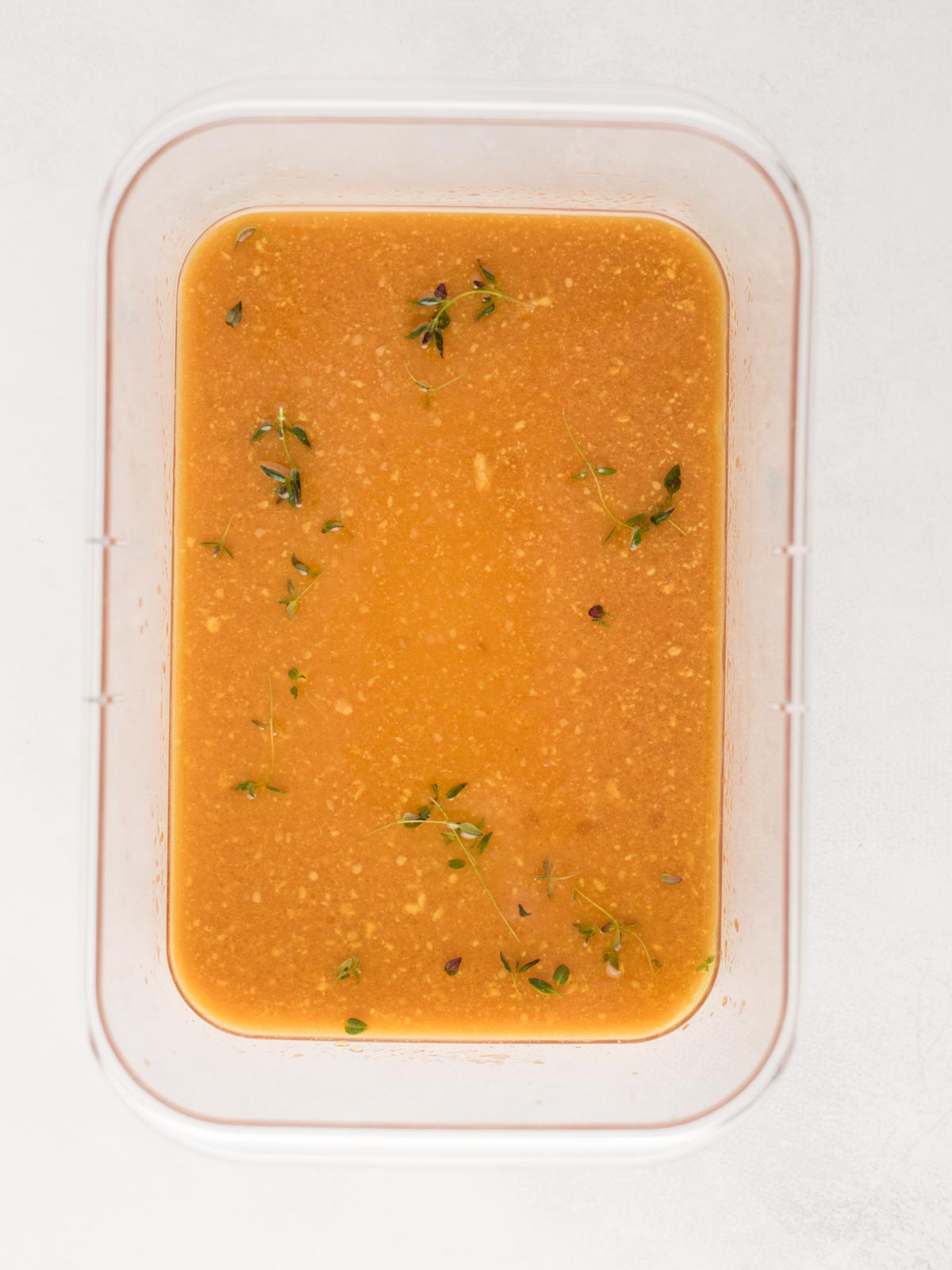 A rectangular container filled with orange-brown marinade, dotted with small bits of herbs and spices for grilled pork chops, with a few sprigs of fresh thyme floating on the surface. The container rests on a white background.