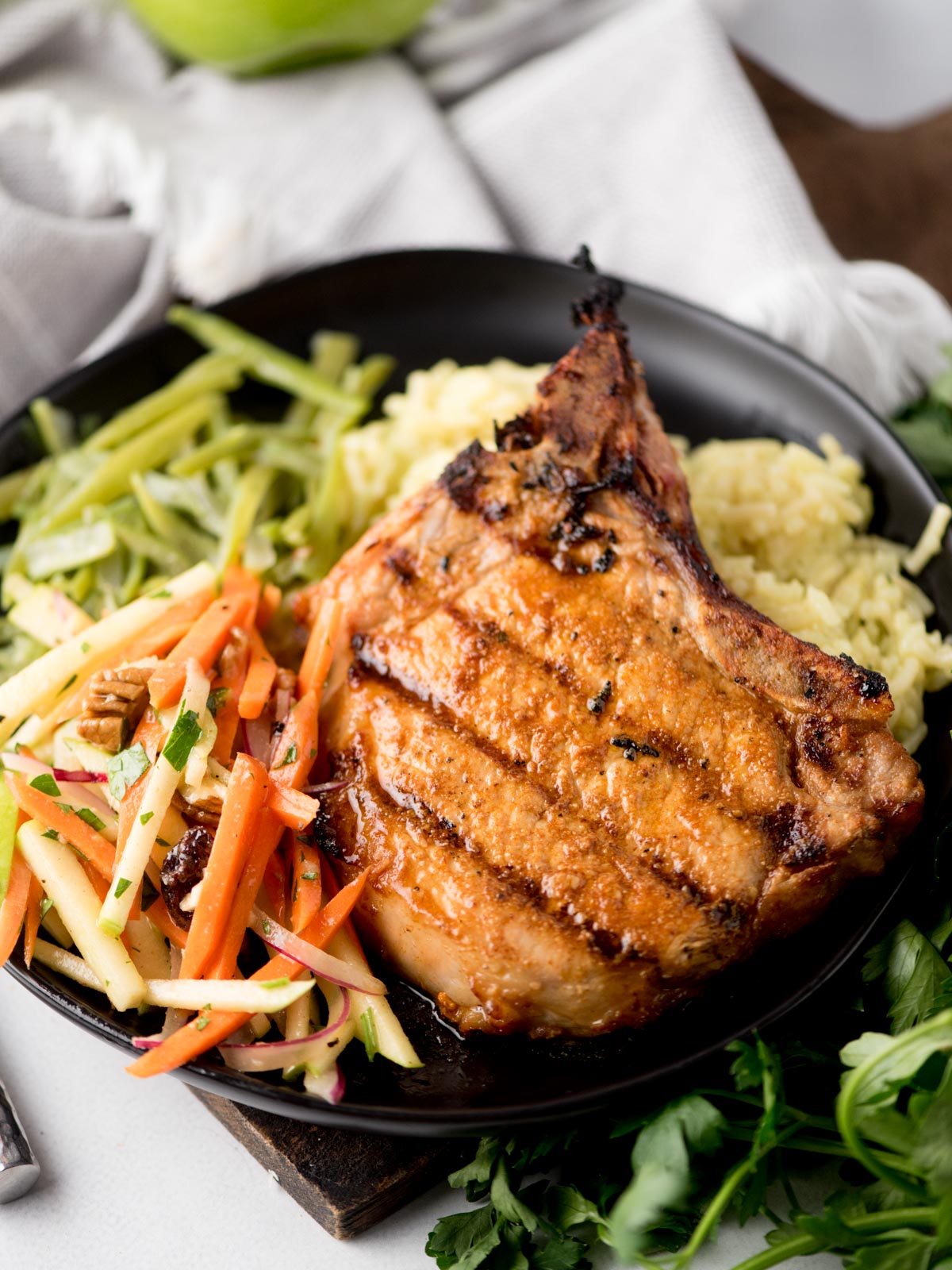 A grilled pork chop served on a black plate with creamy mashed potatoes, crisp green beans, and a colorful apple slaw with julienned carrots and apples, garnished with herbs—a delightful way to enjoy grilled pork chops.