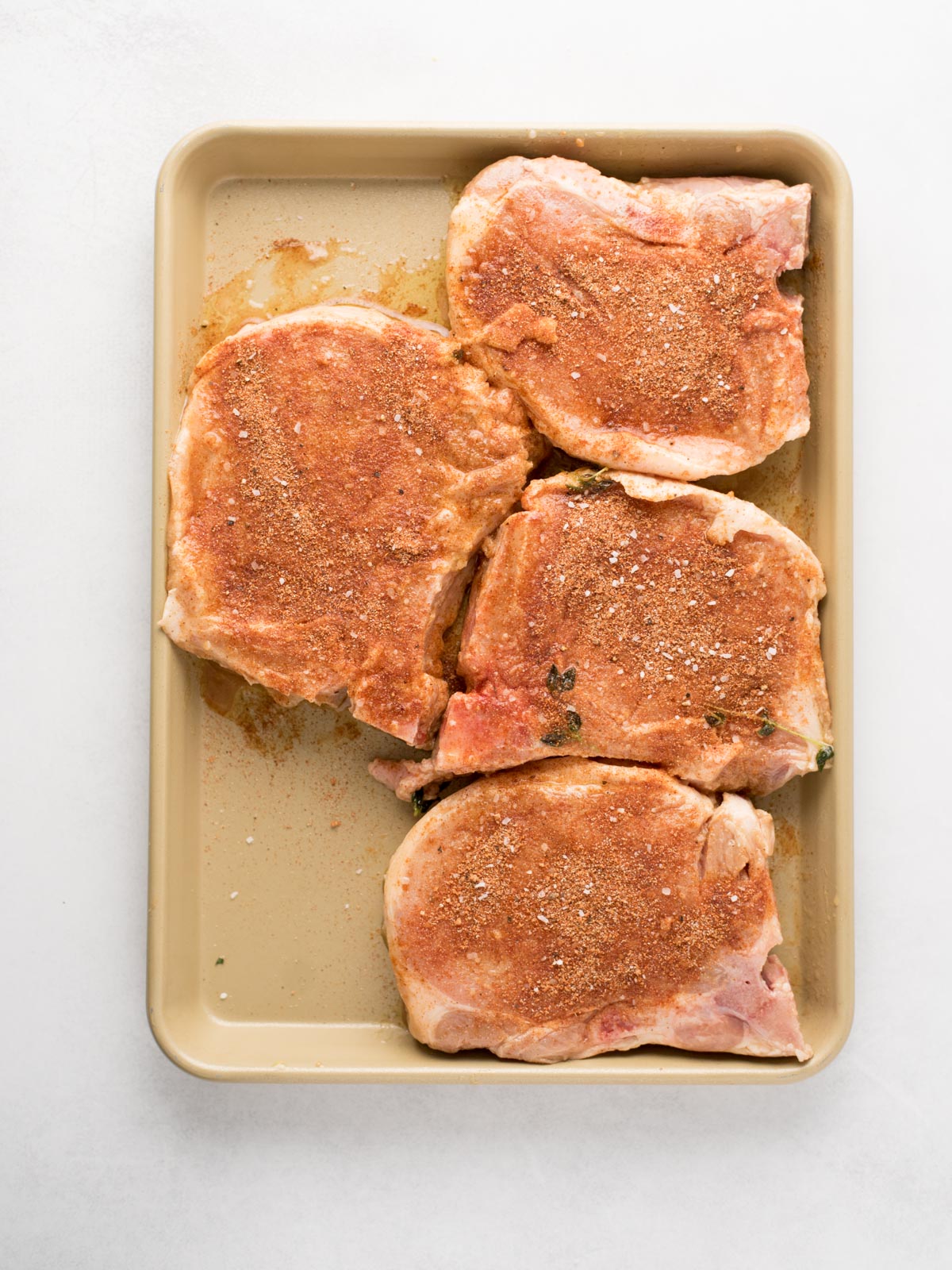 A baking tray with four raw pork chops sprinkled with seasoning, arranged on a light surface—perfectly prepped for delicious grilled pork chops.