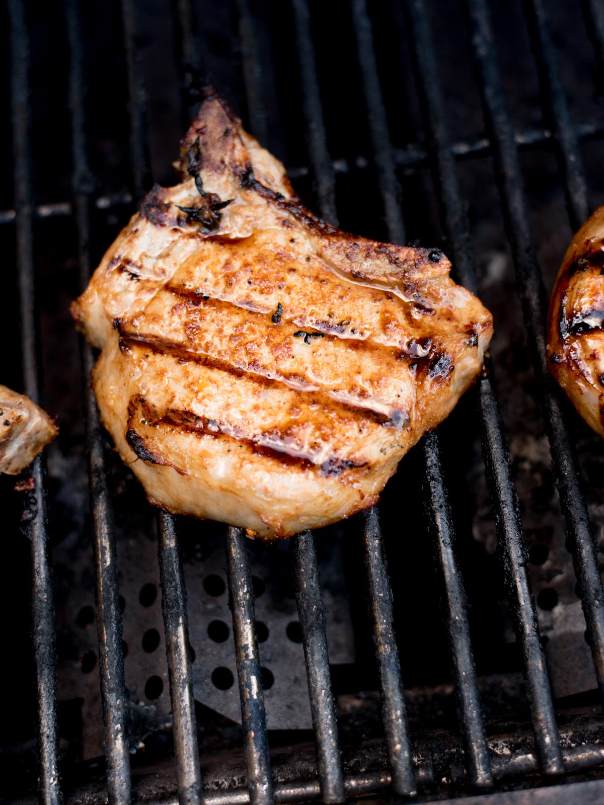 A grilled pork chop with visible grill marks cooks on a metal barbecue grill, showcasing charred lines and a golden-brown exterior—an irresistible example of perfectly grilled pork chops.