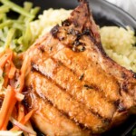 A marinated grilled pork chop served on a black plate with mashed potatoes and a colorful vegetable slaw, garnished with fresh herbs.