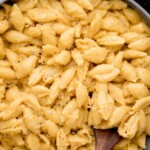 A skillet filled with creamy stovetop mac and cheese made with shell pasta, topped with cracked black pepper. A wooden spoon rests in the pan, and a plate of green beans is partially visible in the upper right corner.