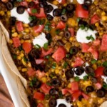 A baked taco casserole in a white dish, topped with olives, tomatoes, green onions, corn, melted cheese, and dollops of sour cream. Text overlay reads weeknight taco casserole.