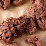 Chocolate cookies with chocolate chips are scattered on brown parchment paper. A cut-out label at the top reads “triple chocolate cookies” above “sweetlysplendid.com” in smaller text.