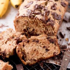 A close-up of sliced banana bread with melted chocolate chunks on top, resting on a cooling rack. Bananas and pieces of chocolate are visible in the background.