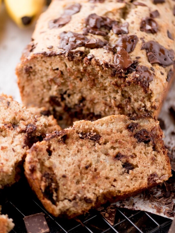 A close-up of sliced banana bread with melted chocolate chunks on top, resting on a cooling rack. Bananas and pieces of chocolate are visible in the background.